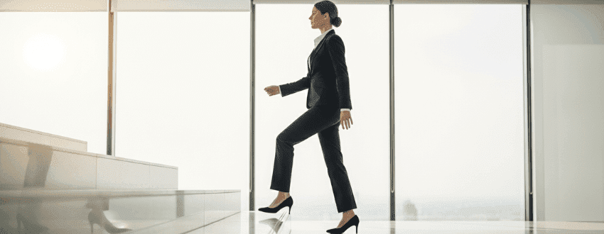 breaking-broken-rung-women-promotion-ladder Professional woman stepping up the career ladder in an office, symbolizing breaking the broken rung and securing first promotion.